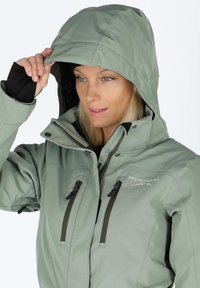 Green softshell jacket with a hood, featuring dual front zip pockets, black inner lining, and contrasting hardware details. "SWEDMOUNT" logo displayed.