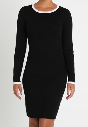 Kobieta ubrana w dopasowaną czarną sukienkę na długi rękaw z białym wykończeniem przy dekolcie i mankietach, stojąca na jednolitym jasnym tle.