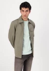Jachetă ușoară de culoare verde măsliniu, cu fermoar frontal și buzunar pe piept, asortată cu o cămașă de culoare verde mentă și blugi de culoare maron închis.