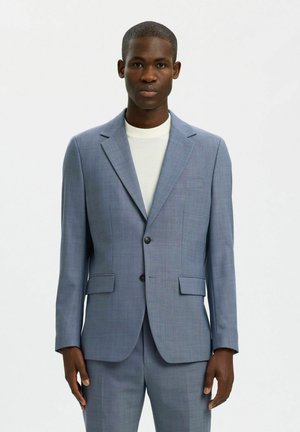 Homme portant un costume bleu clair sur mesure et une chemise blanche à col rond, debout devant un fond blanc uni.