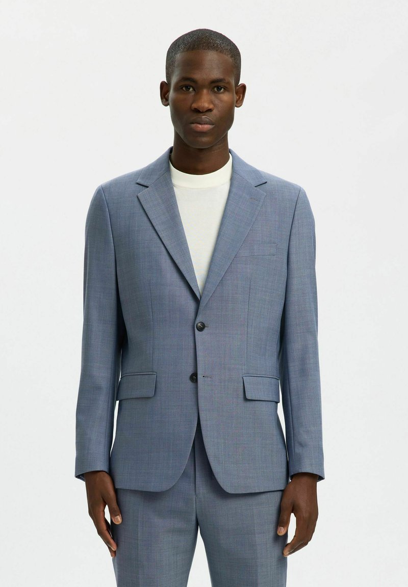 Homme portant un costume bleu clair sur mesure et une chemise blanche à col rond, debout devant un fond blanc uni.