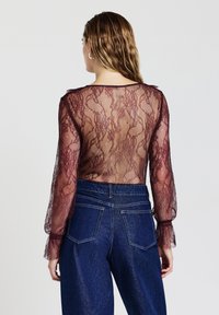 Top in pizzo color bordeaux con maniche lunghe e trasparenti e polsini arricciati; schiena trasparente con motivo in pizzo intricata abbinata a jeans di denim blu a vita alta.