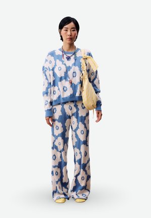 Femme portant un pull et un pantalon à fleurs bleu et blanc, des colliers en perles superposés, un sac jaune sur l'épaule et des chaussures jaunes, debout face à l'avant.
