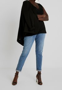 Blazer nero oversize con maniche arrotolate, indossato sopra una maglia nera con pizzo, abbinato a jeans slim-fit azzurri chiari e sandali neri con tacco.