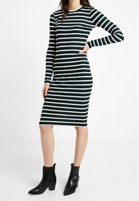 Gestreiftes langärmliges Kleid in Grün und Weiß, aus geripptem Stoff, figurbetonte Form, knielang, kombiniert mit schwarzen Stiefeletten mit Absatz.