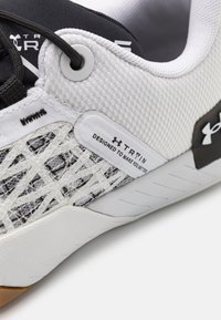 Close-up af hvid og sort Under Armour træningssko med meshstof, snørebånd og etiketten "TRAIN DESIGNED TO MAKE YOU BETTER" på siden.