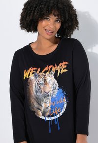 Camisa negra de manga larga con un gráfico de un tigre, salpicaduras de color azul y texto naranja que dice "BIENVENIDO a mi vida salvaje". Textura suave.