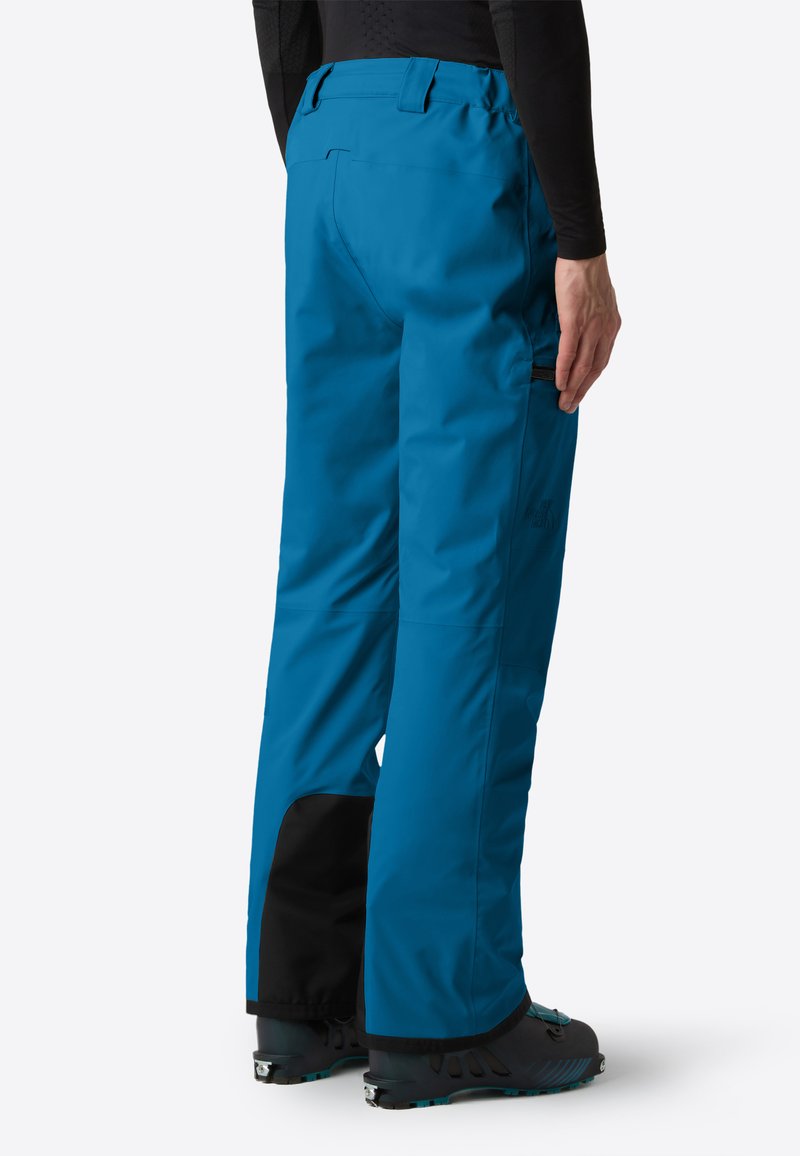 Persona che indossa pantaloni da sci blu con fondo rinforzato nero e scarponi da sci, mostrata di spalle su uno sfondo bianco.