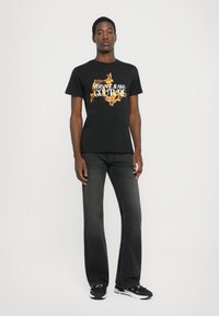 Črna bombažna majica s pisanimi grafičnimi potiski in napisom "VERSACE JEANS COUTURE", v kombinaciji s temno sivo žametnimi kavbojkami in črnimi supergami.