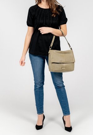 Beige tas met textuur, flap en verstelbare schouderband, gecombineerd met een zwarte top en blauwe spijkerbroek. Vrouw in zwarte hoge hakken staat tegen een effen achtergrond.