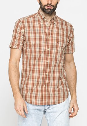 Uomo che indossa una camicia a maniche corte con motivo a quadri marrone e beige e jeans azzurri slavati su sfondo bianco.