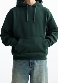 Personne portant un sweat à capuche vert foncé avec une poche avant et un jean bleu, les mains glissées dans la poche, debout devant un fond blanc.