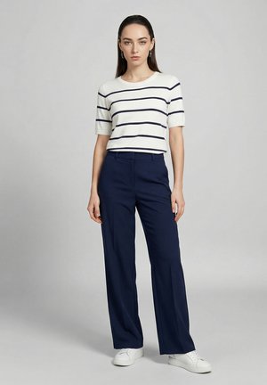 Kvinde står iført hvid kortærmet skjorte med marineblå vandrette striber, marineblå bukser med vide ben, hvide sneakers og hængeøreringe.