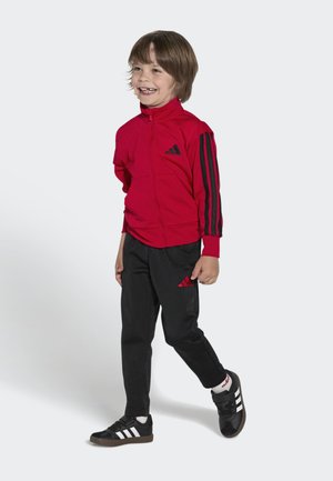 Usměvavý chlapec kráčí, má na sobě červenou bundu na zip s černými pruhy, černé kalhoty s červeným logem, bílé ponožky a černé tenisky Adidas.
