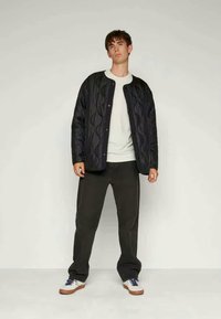 Schwarze gesteppte Jacke mit rundem Kragen, kombiniert mit einem weißen Hemd und dunklen Hosen. Verfügt über Knöpfe und eine strukturierte Oberfläche. Weiße Sneakers.
