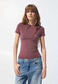 Krátký rukáv, přiléhavý top v barvě vyprahlé růže, s elegantním designem, hladkou texturou a minimem švů. V kombinaci se světle modrými džíny.