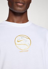 Hvit bomull t-skjorte med rund logo med blomstermønster og gull detaljer. Logoen har teksten "NIKE COURT" under. Kortermet design.