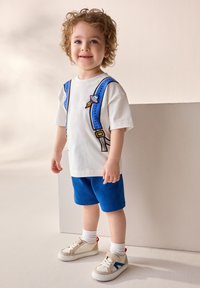 White t-shirt with blue suspenders and space-themed embroidery, paired with blue shorts and white sneakers featuring blue and red accents.