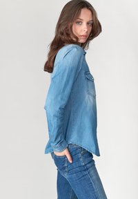 Chemise en denim bleu clair avec des manches longues, un devant à boutons et une poche poitrine unique. Le tissu a une texture douce et une finition légèrement décolorée.