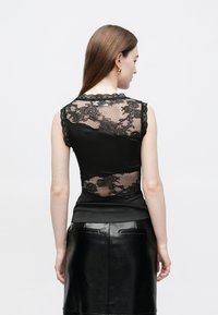 Juodas rankovių neturintis viršus supermatome nėrinių nugara su gėlių raštais ir banguotais kraštais, kartu su lygiu, prilipintu apatiniu skyriumi.