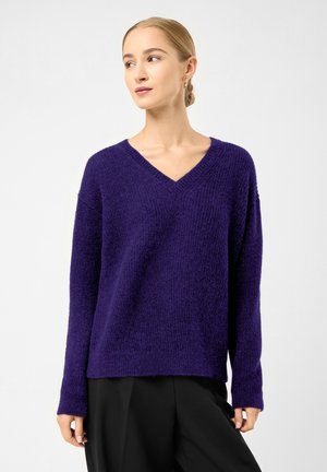 Woman with blonde hair in a low bun wearing a purple V-neck sweater and black pants, looking to the side against a plain light background.
