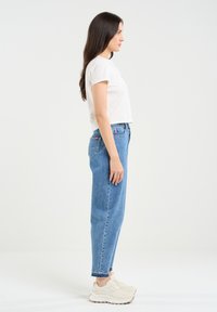 Weißes, kurzes T-Shirt, hellblaue High-Waist-Jeans und beiges Sneakers. Glatter Stoff, lockere Passform, mit sichtbaren Nähten und Taschen.