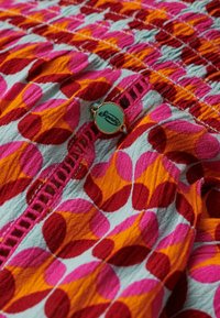 Tela multicolorida con un patrón de formas ovaladas alternadas en rosa, rojo y naranja sobre un fondo azul claro, que presenta una etiqueta metálica de marca.