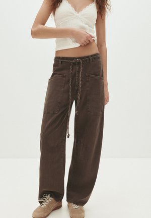 Brown wide-legged pants with two front pockets, a braided drawstring waistband, and a relaxed fit; paired with a light-colored top.