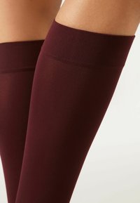 Burgunderfarbene Oberschenkelstrümpfe aus glattem Material, mit einem breiten gerippten oberen Bündchen und nahtlosem Design für eine schlanke Passform.