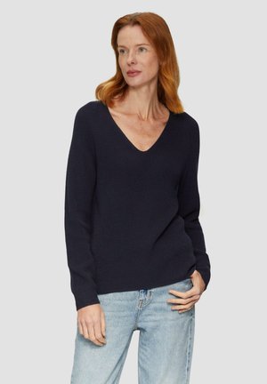Marineblauwe geribde gebreide trui met V-hals en lange mouwen, met een losse pasvorm en subtiele textuur. Gekleed met lichtblauwe jeans.