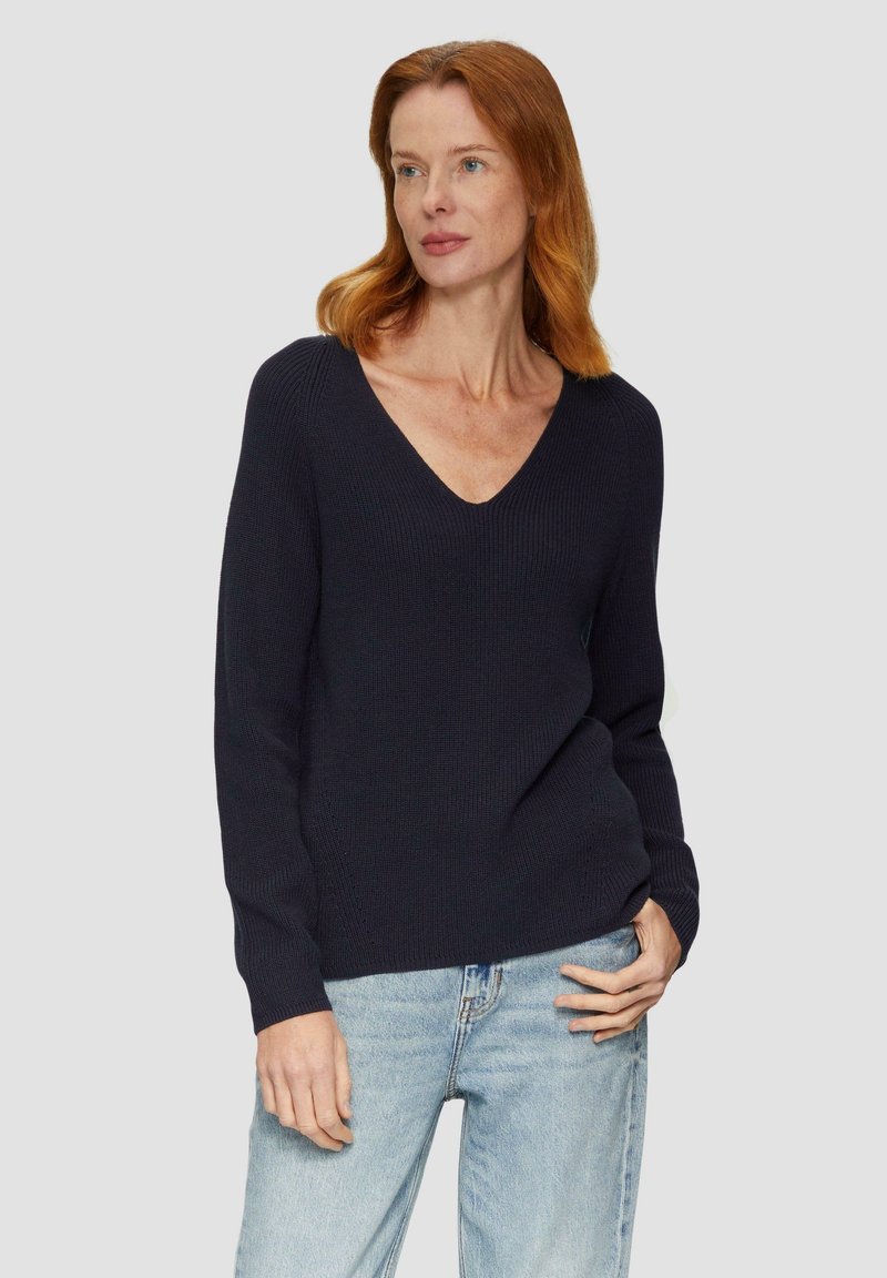 Marineblauer, gerippter Strickpullover mit V-Ausschnitt und langen Ärmeln, in lockerer Passform und mit subtiler Textur. Kombiniert mit hellblauen Jeans.