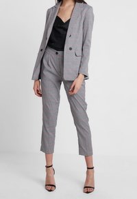 Harmaat ja punaiset ruutukuosiset bleiseri ja housut -setti, jossa on rakenteellinen muotoilu, sileä kangas ja nilkkapituinen lahje. Musta satiinipaita alla.