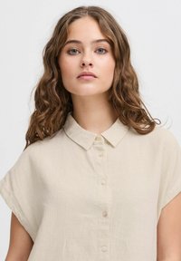 Camicia in lino beige con colletto, maniche corte e chiusura con bottoni. La texture del tessuto sembra leggera e morbida.