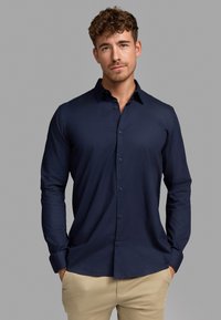 Camisa de botones de manga larga, color azul marino, con cuello, textura suave, un bolsillo en el pecho y botones a juego. Llevada con pantalones de color claro.