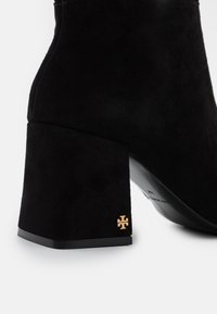 Bottine en daim noir avec talon bloc. Comprend un détail logo doré sur le talon, une texture très douce et un design épuré et minimaliste.