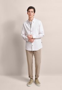 Weißes Langarmhemd mit Knöpfen vorne, leichtes Material und Stehkragen kombiniert mit beigen Chinos und beigen Sneakers.