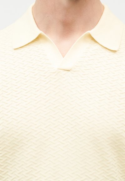 Κοντινό πλάνο ενός ατόμου που φοράει ένα ελαφρώς μπεζ πόλο με υφή, με διπλωμένο γιακά και λαιμόκοψη σε σχήμα V.