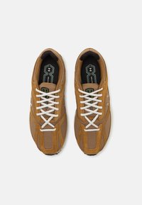 Brune sneakers lavet af mesh og ruskindsmaterialer, med hvide snørebånd og en struktureret sål. Logoet er trykt i en kontrastfarve.