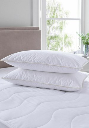 Deux oreillers blancs empilés sur un matelas blanc matelassé dans une chambre lumineuse avec une fenêtre et des plantes vertes en arrière-plan.