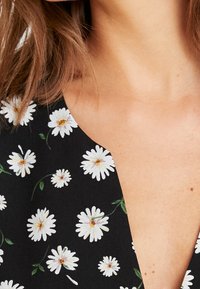 Tissu noir avec motif floral de marguerites blanches, présentant un col en V profond et une texture légère. Accents verts parmi les fleurs.