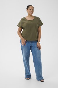 Femme souriante, debout avec les mains détendues, portant un haut à manches courtes vert olive, un jean bleu à jambes larges, des sandales noires à bout ouvert et des bijoux en argent.