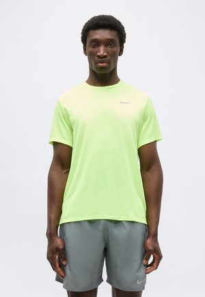 Homme en t-shirt Nike vert clair et short Nike gris, debout devant un fond uni, face à l'avant avec une expression neutre.