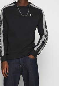 Black sweatshirt with raglan gray sleeves, featuring logo on the chest and graphic accents along the sleeves. Smooth texture and fitted design.