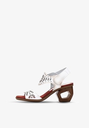 Sandale en cuir blanc avec des motifs découpés, semelle en daim rouge et un talon en bois hexagonal unique. Les brides maintiennent le pied avec un design minimal.