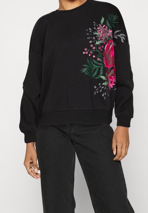 Sort langærmet sweatshirt med pink og grøn blomstret print på venstre side, båret med sorte jeans.
