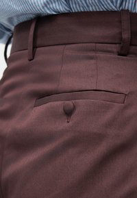 Κοντινό πλάνο στην τσέπη παντελονιού μπορντό με κλείσιμο με κουμπί, ραμμένους θηλιές ζώνης και μια ριγέ μπλε πουκάμισο κρυμμένη μέσα.