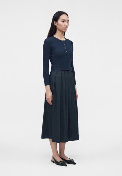 Femme debout portant un cardigan bleu marine ajusté boutonné, une jupe midi plissée taille haute et des ballerines slingback noires pointues.
