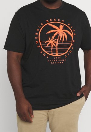 Mężczyzna ubrany w czarną koszulkę z pomarańczowym nadrukiem palmy i napisem "The Emerald Beach Club Resort 1969", w połączeniu z beżowymi spodniami.