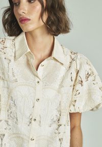 Weißes Blusenshirt mit aufwändigen Spitzenverzierungen, kurzen Puffärmeln und einem spitzen Kragen. Knöpfe entlang der Vorderseite verleihen dem Design eine besondere Note.