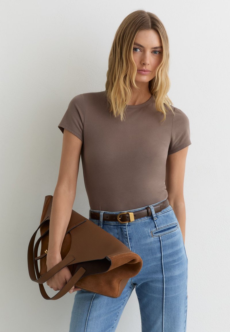 Femme aux cheveux blonds portant un haut marron à manches courtes, un jean bleu taille haute et tenant un grand sac à main en cuir marron.